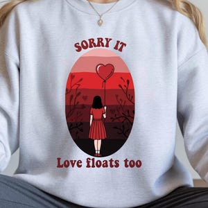 Könnte beinhalten: Ein hellgraues Sweatshirt mit dem Text "Sorry it" über "Love floats too". Die Grafik zeigt ein Mädchen, das einen herzförmigen Ballon vor einem roten und kastanienbraunen Sonnenuntergang hält. Das Mädchen trägt ein rotes Kleid.