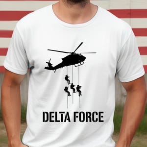 May include: White t-shirt featuring a black graphic of a helicopter with soldiers rappelling down. The text "DELTA FORCE" is printed in bold, black letters below the image. The background includes an American flag.