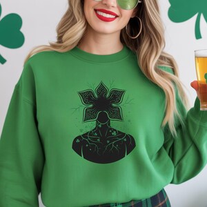 May include: Green sweatshirt featuring a black graphic of a floral-like head and a silhouette of a torso with shamrock accents. The sweatshirt is a solid green color. The background has shamrock decorations.