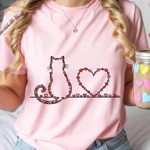 May include: Pink t-shirt featuring a cat silhouette and a heart, both outlined with red hearts. The cat sits beside a heart shape, also made of red hearts, on a black line. A jar with heart-shaped stickers is visible.