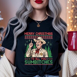 May include: Black t-shirt with a Christmas-themed graphic. The design features a cartoon sheriff with a cowboy hat and the text "MERRY CHRISTMAS TO ALL YOU SUMBITCHES" in red and green. The shirt has a festive, humorous style.