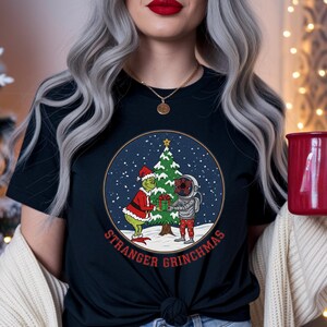 Puede incluir: Camiseta negra con un gráfico circular que presenta al Grinch con un traje de Papá Noel regalando un obsequio a un astronauta con un traje espacial. Un árbol de Navidad está en el fondo. El texto "STRANGER GRINCHMAS" está impreso debajo.