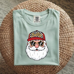 May include: A light green t-shirt featuring a graphic of Santa Claus wearing a red cap with a checkered pattern and the word "Jolly". Santa is also wearing star-shaped sunglasses. The shirt is folded on a woven surface.