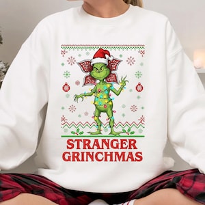 Puede incluir: Sudadera blanca con una criatura verde con gorro de Papá Noel y luces navideñas. El diseño incluye detalles en rojo y verde con copos de nieve y el texto "STRANGER GRINCHMAS" en rojo.
