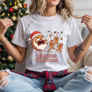 Könnte beinhalten: Weißes T-Shirt mit einem weihnachtlichen Motiv. Das Design zeigt ein Weihnachtsmann-Gesicht aus einem Keks, Lebkuchenmännern, Keksen und Zuckerstangen. Der Text "DELICIOUS CHRISTMAS" ist darunter gedruckt.