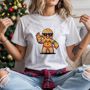 Könnte beinhalten: Weißes T-Shirt mit einem Cartoon-Lebkuchenmann, der eine Sonnenbrille trägt und eine Zigarette raucht. Der Lebkuchenmann ist mit Zuckerguss und festlichen Akzenten verziert. Das T-Shirt wird von einer Person in blauen Jeans getragen.