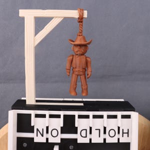 May include: A brown cowboy figurine hangs from a wooden gallows. The gallows is made of light brown wood. The base of the item has the words "HOLD ON" in black and white letters.