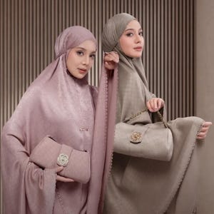 May include: Two women wearing matching headscarves and robes, each holding a purse. The robes are in shades of mauve and taupe. The purses have a floral clasp and a gold chain strap.