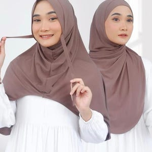 Puede incluir: Dos mujeres con hiyabs marrones. Los hiyabs están drapeados sobre la cabeza y los hombros, con un detalle de lazo. Las mujeres visten vestidos blancos. El fondo es blanco liso.