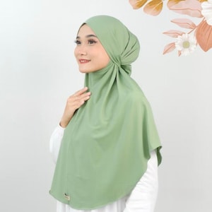 May include: A sage green hijab featuring a smooth, opaque fabric that drapes over the head and shoulders. The head covering is styled with a fitted cap section and a flowing, rounded front panel that extends down to the chest area, creating a clean look.