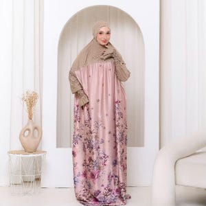 Könnte beinhalten: Ein bodenlanges, locker geschnittenes Kleid in zartem Rosa mit einem Blumenmuster in Violett- und dunklen Farbtönen. Das Kleidungsstück umfasst eine passende beige Kopfbedeckung und lange Ärmel mit gerafften, strukturierten Bündchen.