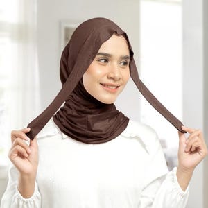 May include: A dark brown hijab featuring a smooth, fitted hood that covers the head and neck, with two long, flat fabric ties extending from the sides. The fabric has a soft, matte finish and drapes gently around the neck area in subtle folds.