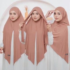 May include: Three women wearing matching peach-colored hijabs. The hijabs are draped over the heads and shoulders, with a long, flowing front panel. The women are wearing white tops. The hijabs appear to be made of a soft, lightweight fabric.