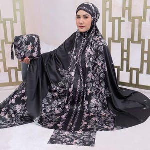 May include: A black prayer garment featuring a floral pattern in shades of pink, white, and grey. The set includes a matching head covering and a small rectangular pouch with a wrist strap, both decorated with the same intricate botanical print.