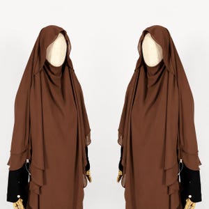 Può includere: Un khimar marrone caratterizzato da un design a strati con cappuccio drappeggiato e tessuto fluido che scende fino al busto. L'indumento presenta piccoli dettagli color argento sulle balze laterali ed è esposto su un manichino con maniche nere.