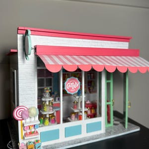 May include: Miniature candy shop with a white brick facade, pink and white striped awning, and a sign that reads "Candy Shop." The shop features shelves filled with colorful candies and lollipops. A whimsical and detailed model.