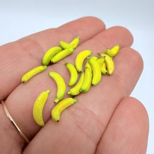 Pode incluir: Miniaturas de bananas amarelas com pontas verdes estão espalhadas sobre uma mão. As bananas são pequenas, curvas e parecem ser feitas de um material liso. O fundo é branco liso.