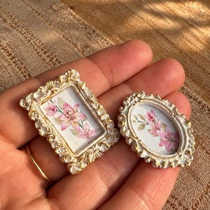 Puede incluir: Dos marcos de fotos en miniatura con bordes ornamentados en blanco y oro con acabado desgastado. Un marco es rectangular y el otro es ovalado. Ambos contienen una pequeña ilustración centrada de flores de lirio rosas con hojas verdes sobre fondo blanco.