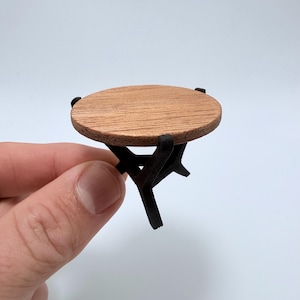 Könnte beinhalten: Ein Miniatur-Rundtisch mit hellbrauner Holzplatte und schwarzen Beinen. Der Tisch wird in einer Hand vor weißem Hintergrund gehalten. Die Tischplatte hat einen Durchmesser von ca. 5 cm.