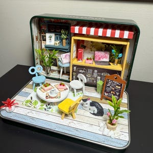 Könnte beinhalten: Miniatur-Diorama eines Cafés in einer grünen Dose. Enthält ein Café mit rot-weiß gestreifter Markise, kleine Tische, Stühle und eine Menütafel. Enthält winzige Pflanzen, Desserts und eine Katzenfigur.