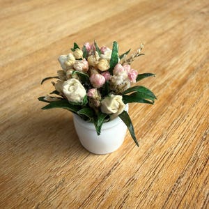 Op de afbeelding: Een miniatuur bloemstuk met kleine witte en roze gedroogde bloemen en groene bladeren, geplaatst in een eenvoudige, gladde witte keramische pot. Het boeket is dicht gevuld met delicate bloesems en bladeren voor een compact geheel.
