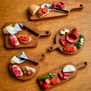 May include: Five miniature wooden serving boards arranged on a surface, each displaying tiny food items including slices of bread, red tomatoes, green pickles, cured meats, white cheese, and small black knives with silver blades and foil wrappers.