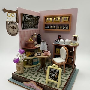 Puede incluir: Diorama en miniatura de una cafetería con una pared rosa, una pizarra de menú y una pequeña mesa con una taza rosa. La escena incluye un molinillo de café, una silla pequeña y un letrero que dice "Coffee Corner".