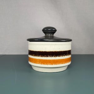 Peut inclure: Un sucrier en céramique avec un couvercle noir et une base blanche. Le bol présente des rayures horizontales marron et orange. Le couvercle a un bouton arrondi. Le bol est conçu pour la cuisine.