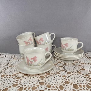 May include: A set of six cream-colored teacups and saucers. Each cup and saucer features a pink floral design with gray and yellow accents. The cups are stacked and arranged on a white lace tablecloth.