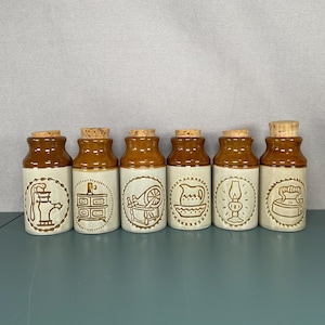 May include: A set of six vintage ceramic jars with cork stoppers. The jars have a brown top and a cream base. Each jar features a different embossed design, including a teapot, dresser, spinning wheel, bowl, lamp, and iron.