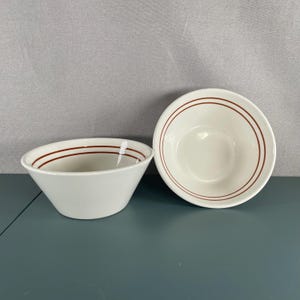 Puede incluir: Dos cuencos de cerámica de color blanco roto con dos finas rayas paralelas de color óxido que rodean el borde interior. Un cuenco está colocado en ángulo mostrando el perfil lateral, mientras que el otro está en posición vertical.