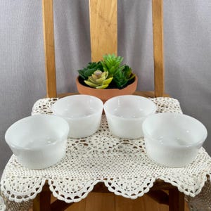 May include: Four white, round, textured bowls are arranged on a cream-colored, lace-trimmed cloth. A small terracotta pot with a succulent plant sits in the background. The bowls are on a wooden chair.