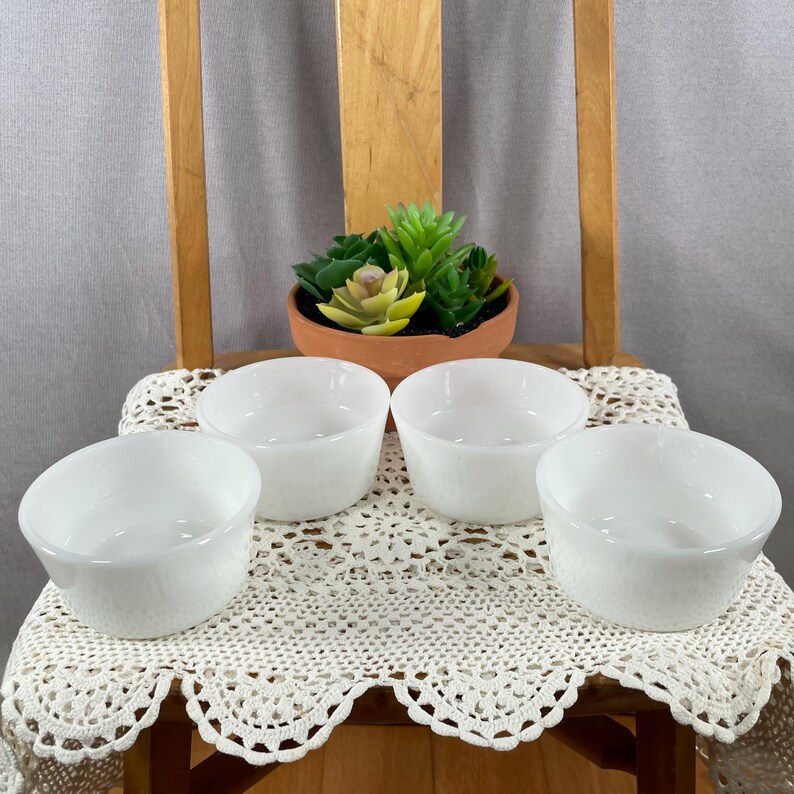 May include: Four white, round, textured bowls are arranged on a cream-colored, lace-trimmed cloth. A small terracotta pot with a succulent plant sits in the background. The bowls are on a wooden chair.