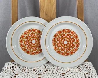 May include: Two white ceramic plates with an orange and yellow floral design in the center. Each plate has a thin orange border. The plates are displayed on a wooden chair.