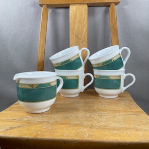 May include: A set of white ceramic teacups and a creamer with a teal and gold geometric design. The cups are stacked on a wooden chair. The creamer has a spout for easy pouring.