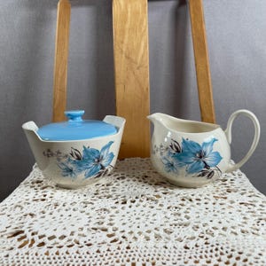 May include: A vintage cream and sugar set. The sugar bowl has a light blue lid and handles. Both pieces are decorated with blue floral designs on a white background. The set is displayed on a white lace doily.