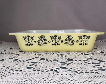May include: A rectangular, vintage Pyrex baking dish with a pale yellow exterior and white interior. The dish features a black floral pattern around the sides. The dish has a textured exterior and a handle on each end.