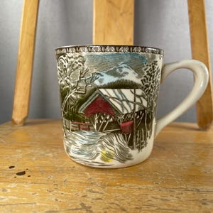 May include: A cream-coloured ceramic mug featuring a detailed illustration of a covered bridge over a stream, surrounded by trees and mountains. The design includes a decorative border at the top rim and a sturdy handle on the right side.