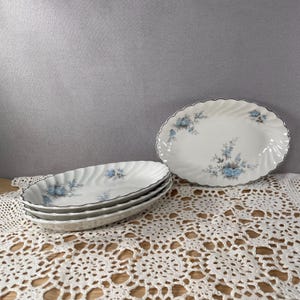 Op de afbeelding: Een set witte ovale serveerschalen met een blauw bloemmotief en een grijze rand. De schalen zijn gestapeld en gerangschikt op een wit kanten tafelkleed. Eén schaal is apart geplaatst.