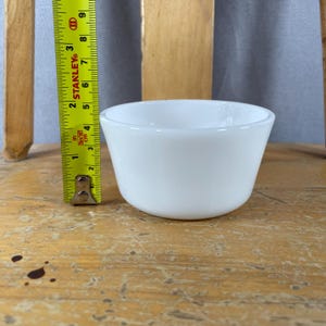 May include: A small, white, opaque glass bowl. The bowl is approximately 3 inches in diameter and 2 inches tall. The bowl is sitting on a wooden surface. A yellow measuring tape is next to the bowl.
