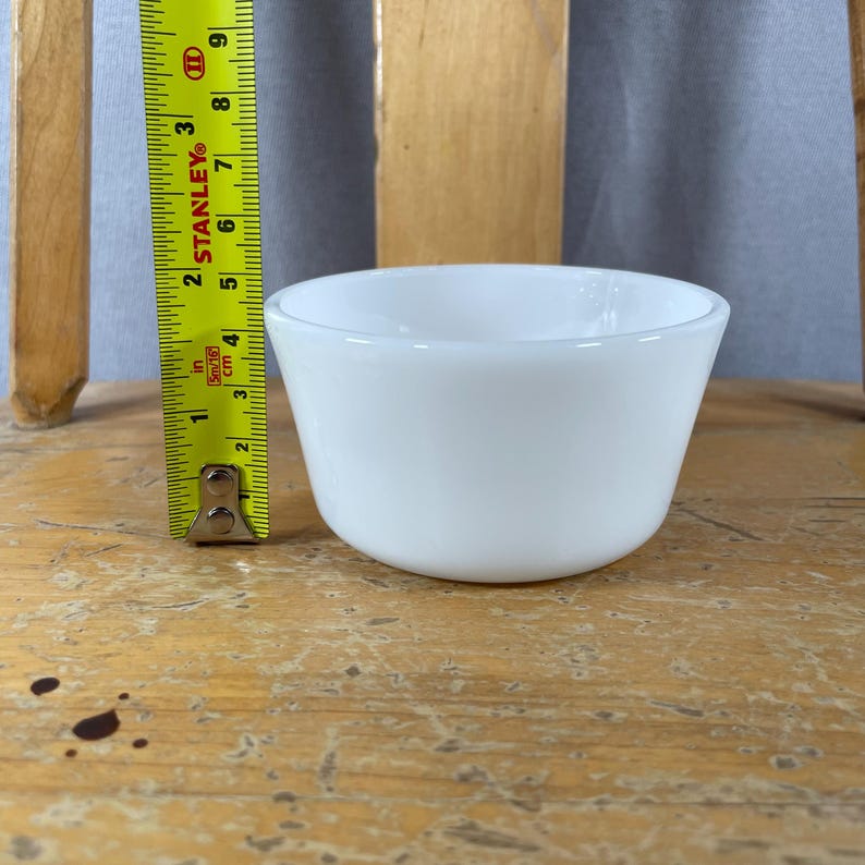 May include: A small, white, opaque glass bowl. The bowl is approximately 3 inches in diameter and 2 inches tall. The bowl is sitting on a wooden surface. A yellow measuring tape is next to the bowl.