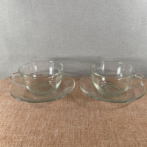 May include: A pair of clear glass teacups and saucers. The cups have a rounded design with a handle and sit on clear, circular saucers. The set is made of transparent glass. The background is a neutral colour.