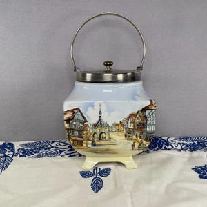 May include: A vintage ceramic jar with a silver lid and handle. The jar features a painted scene of a town with buildings and a church. The jar has a light blue sky background and sits on a white surface.