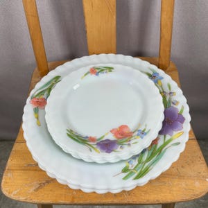 May include: A stack of three white ceramic plates with scalloped edges. Each plate is decorated with a floral design featuring pink, purple, and blue flowers with green stems and leaves. The plates are stacked on a wooden chair.