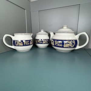 May include: A white ceramic tea set consisting of a teapot, a lidded sugar bowl, and a creamer. Each piece features a wide navy blue band decorated with gold filigree patterns and the text Tetley Teas in gold lettering, accented by thin silver rims.