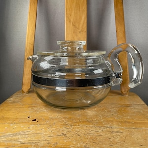 May include: A clear glass teapot with a rounded body and a matching glass lid. The teapot features a wide, horizontal metallic band encircling the centre and a sturdy, curved glass handle attached to the side with a visible metal screw fixture.