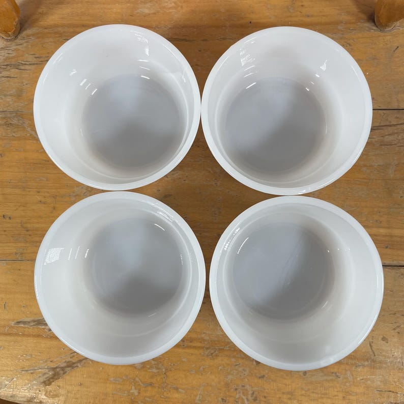 May include: Four white, round bowls are arranged on a wooden surface. The bowls have a simple, classic design with a smooth, glossy finish. The bowls are empty and ready for use.