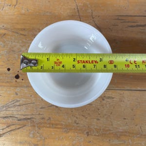 May include: A white, round bowl with a 3-inch diameter is measured with a yellow Stanley tape measure. The bowl is placed on a wooden surface.