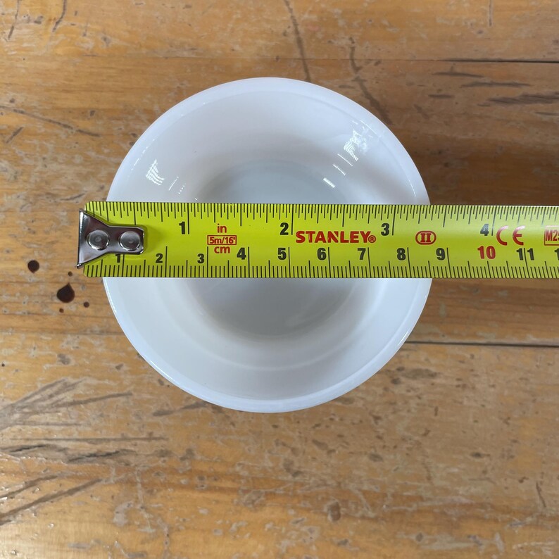 May include: A white, round bowl with a 3-inch diameter is measured with a yellow Stanley tape measure. The bowl is placed on a wooden surface.