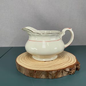 Peut inclure: Crémier en céramique crème avec un bord doré et une ligne rouge. Le crémier a une anse et un bec verseur, et repose sur une tranche de bois. Une pièce classique pour servir la crème ou les sauces.